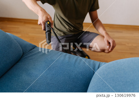 Cropped shot of cleaning service worker using steam cleaner to remove dirt and stains from blue sofa in modern living room, ensuring hygiene and freshness. Concept of easy housekeeping domestic life. 130619976