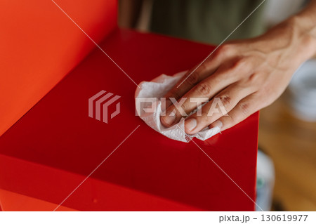 Male hand wiping dry red surface of shelf with white paper towel after housekeeping at home, close-up. Concept of cleanliness and hygiene in domestic environment. 130619977