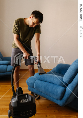 Vertical portrait of young man using washing vacuum cleaner to clean blue sofa in home setting, demonstrating modern approach to household chores and upholstery maintenance. 130619978