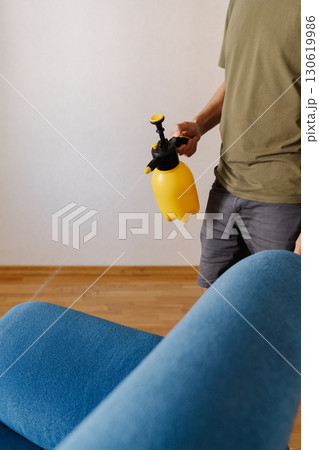 Househusband spraying water onto blue sofa using yellow sprayer, diligently cleaning furniture in cozy living room at home, ensuring hygiene and freshness, close-up. Concept of hygiene and cleanliness 130619986