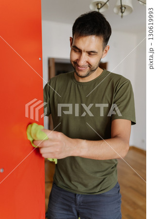 Cheerful young man cleaning red shelf with rag, ensuring dust-free and spotless surface for books and decorative items, enhancing organization and hygiene at home. Concept of household cleanliness. Cheerful young man cleaning red shelf with rag, ensuring dust-free and spotless surface for books and decorative items, enhancing organization and hygiene at home. Concept of household cleanliness. 130619993