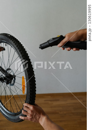 Vertical cropped shot of owner bike cleaning dirty dusty tire of bicycle wheel using steam cleaner, ensuring thorough cleaning in apartment environment, close-up. Concept of bike maintenance, service. 130619995