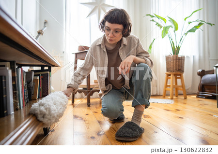 Tidy neat young woman using duster to remove dust from furniture near floor, home cleaning routine 130622832