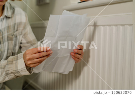 Woman holding utility paper bills next to heating thermostat, increasing heating costs Woman holding utility paper bills next to heating thermostat, increasing heating costs 130623070