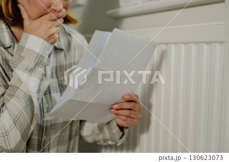 Upset woman holding utility paper bills next to heating thermostat, increasing heating costs Upset woman holding utility paper bills next to heating thermostat, increasing heating costs 130623073