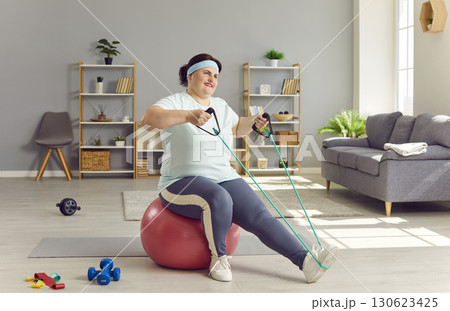 Overweight woman sitting on fit ball at home and doing exercise with resistance band 130623425