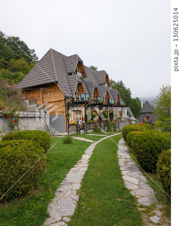 Row of small wooden cottages with pointed roofs, surrounded by trimmed bushes and stone path. Tourism and mountain accommodation in Serbia. 130625014