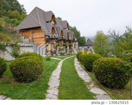 Row of small wooden cottages with pointed roofs, surrounded by trimmed bushes and stone path. Tourism and mountain accommodation in Serbia. 130625015