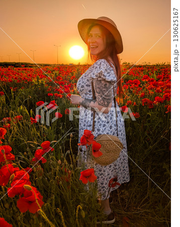 A woman in a floral dress and straw hat stands among poppies at sunset. Lifestyle, identity, and natural connection with rural environment and human individuality. 130625167
