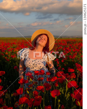 A woman smiles in a straw hat surrounded by red poppies at sunset. Lifestyle, beauty, and natural harmony of rural landscape with human presence and joy. 130625171
