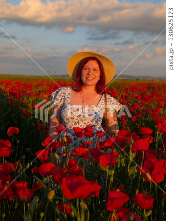 A woman in a floral dress and straw hat stands among poppies at sunset. Lifestyle, identity, and natural connection with rural environment and human individuality. 130625173