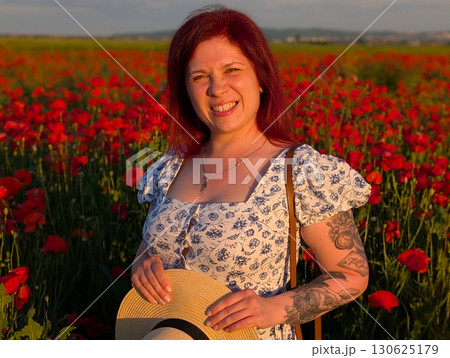 A woman smiles brightly in a poppy field with sunset light on her face. Portrait of happiness, individuality, and emotional lifestyle connection with summer nature. 130625179