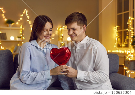 Couple sitting on sofa looking at red heart in bedroom with garlands Couple sitting on sofa looking at red heart in bedroom with garlands 130625304