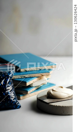 A stack of four square blue tiles with adhesive in between, next to a sealed bag of small blue objects, atop a gray surface; nearby lies a stone with a swirl of beige paste 130626424