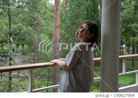 A woman in a casual grey t-shirt stands on a balcony in a green forest in Peredelkino, turning her head to look at the camera 130627009