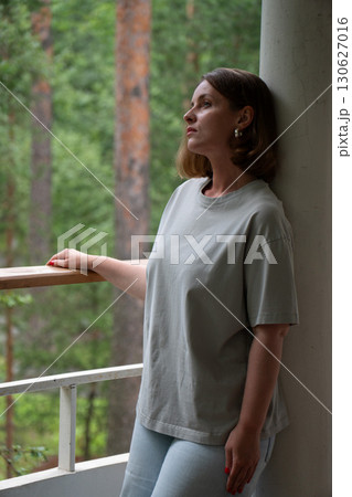 A young woman stands still on a forest balcony in Peredelkino, leaning against a column and looking up with a calm, distant gaze A young woman stands still on a forest balcony in Peredelkino, leaning against a column and looking up with a calm, distant gaze 130627016