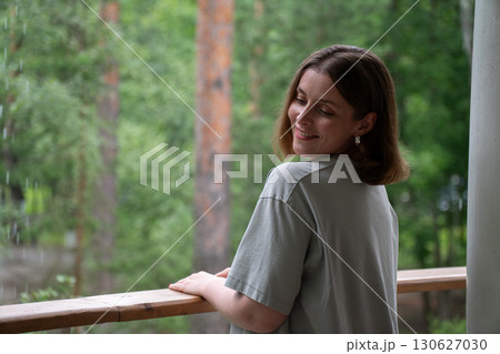 A smiling young woman in a light t-shirt stands on a balcony in the forest in Peredelkino, closing her eyes and enjoying the moment 130627030