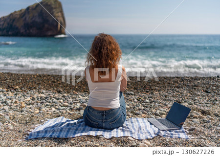 Beach Laptop Woman: Back view of woman using laptop on pebble beach, enjoying coastal views in daytime. Beach Laptop Woman: Back view of woman using laptop on pebble beach, enjoying coastal views in daytime. 130627226