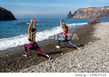 Yoga Beach Exercise: Women stretch on pebble beach by ocean, sunny day, for health and relaxation. Yoga Beach Exercise: Women stretch on pebble beach by ocean, sunny day, for health and relaxation. 130627228