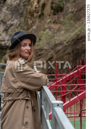 A stylish woman in a beige trench coat and hat stands near a red railing in a rocky canyon, looking thoughtfully into the distance A stylish woman in a beige trench coat and hat stands near a red railing in a rocky canyon, looking thoughtfully into the distance 130627370