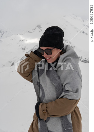 A woman in a beanie, scarf and trench coat smiles gently while standing in a snowy mountain landscape. 130627953