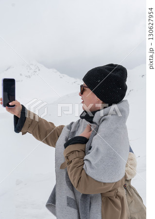 A woman in a winter outfit takes a selfie on her smartphone in a snowy mountain landscape 130627954
