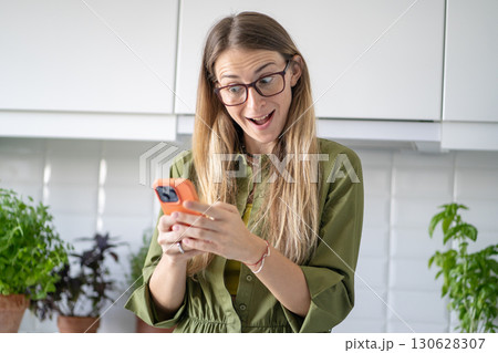 Amazed woman with smartphone receiving good news. Joyful female chatting and video calling at home Amazed woman with smartphone receiving good news. Joyful female chatting and video calling at home 130628307