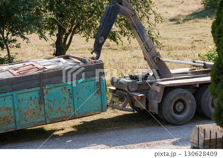 Rusty Dumpster Being Lifted by Crane Truck on Asphalt Roadside with Green Field Backdrop 130628409