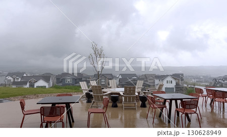 A backyard patio with outdoor furniture is shown under gray skies during a rainstorm. Wet surfaces and distant suburban homes are visible in the background. A backyard patio with outdoor furniture is shown under gray skies during a rainstorm. Wet surfaces and distant suburban homes are visible in the background. 130628949