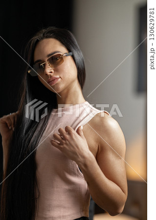 girl with long hair and glasses stands indoors exuding self assurance. She gently adjusts her clothing highlighting her strong physique. The setting has a modern feel with soft lighting. girl with long hair and glasses stands indoors exuding self assurance. She gently adjusts her clothing highlighting her strong physique. The setting has a modern feel with soft lighting. 130629781