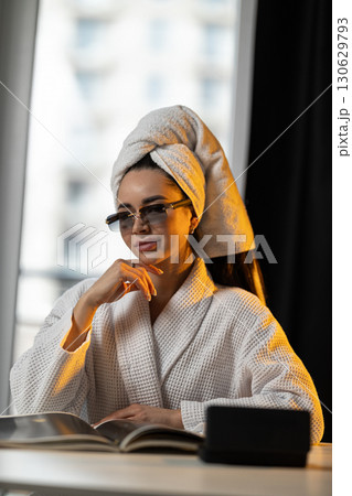 young girl sits comfortably in the kitchen after taking a shower. She wears a towel on her head and glasses basking in the gentle light of dawn as she reflects on her morning. young girl sits comfortably in the kitchen after taking a shower. She wears a towel on her head and glasses basking in the gentle light of dawn as she reflects on her morning. 130629793