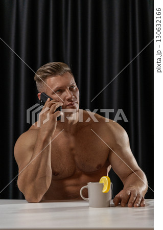 An athletic man sits shirtless at a table talking on the phone with a mug of tea beside him. A slice of lemon decorates the mug creating a warm atmosphere. 130632166