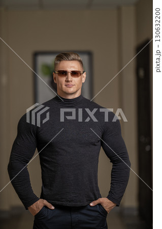 athletic man with glasses stands confidently in a modern indoor setting. His muscular build is emphasized by a fitted black shirt. The environment exhibits clean lines and neutral tones. 130632200