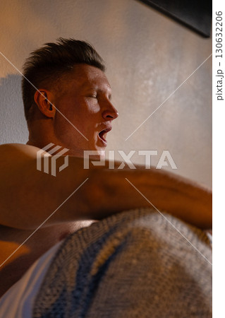athletic man is seen in bed stretching and enjoying a moment of relaxation. The soft light highlights his focused expression as he begins his day. 130632206