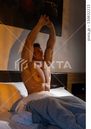Muscular man sits up in bed stretching his arms above his head. Sunlight filters through the window creating a warm atmosphere in the bedroom. 130632215
