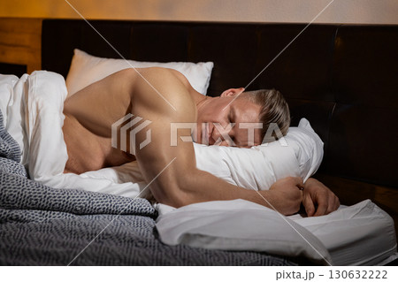 athletic man rests soundly in bed his muscular build relaxed against soft pillows and blankets. The warm lighting and cozy atmosphere indicate a calm end to a strenuous day. athletic man rests soundly in bed his muscular build relaxed against soft pillows and blankets. The warm lighting and cozy atmosphere indicate a calm end to a strenuous day. 130632222