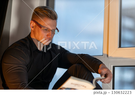 An athletic man with glasses is seated on a windowsill immersed in a book. The soft glow from outside enhances the cozy atmosphere as he reads during the evening. 130632223
