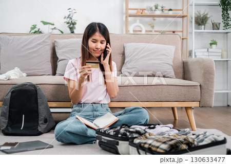 Woman sitting on floor packing suitcase, using phone and credit card, preparing for travel in a cozy living room. Woman sitting on floor packing suitcase, using phone and credit card, preparing for travel in a cozy living room. 130633477