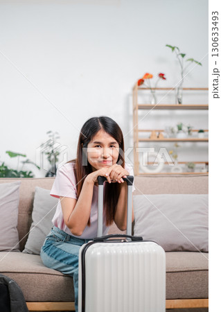 Smiling woman sitting on a sofa with a suitcase, ready for travel. Modern living room ambiance. Smiling woman sitting on a sofa with a suitcase, ready for travel. Modern living room ambiance. 130633493
