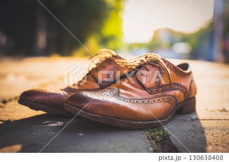 Pair of vintage brown leather brogues with decorative perforations resting on sunny pavement, worn leather showing scuffs and character, classic footwear in warm evening light scene 130638408