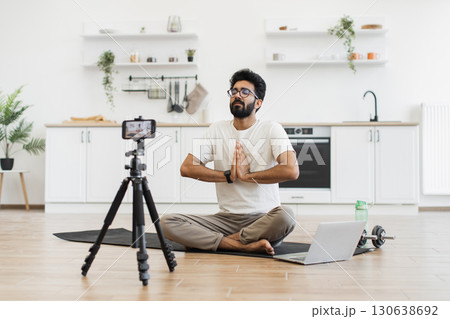 Young adult man sitting in modern kitchen recording video blog about fitness and mindfulness while meditating. Promoting healthy lifestyle including mental and physical wellness to his online audience 130638692