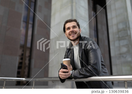 an adult man with a smile stands with a cup of coffee and a mobile phone and looks up 130639836