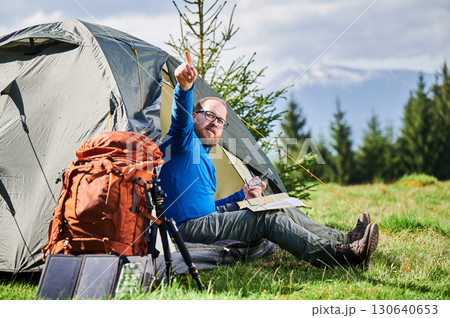 Man sits in tourist tent's entrance, points into distance while studying map. Beside him orange backpack, equipped with tripod and solar panel, set against scenic background of trees and mountains. 130640653