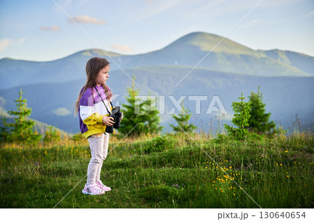 Young girl walks through grassy meadow, holding binoculars, wearing colorful sweatshirt. Lush green hills and distant mountains under clear evening sky, creating serene and picturesque scene. 130640654