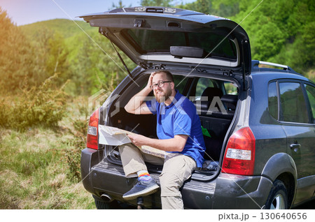 Man sits on open trunk of SUV in scenic, green landscape, holding map and scratching his head in confusion. Wearing blue shirt, beige pants, and colorful socks, he trying to figure out his location. 130640656