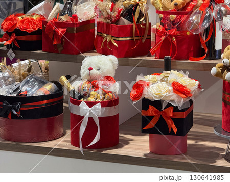 Gift boxes with teddy bears, roses and wine bottles are arranged in a festive display. Romance, love, consumer culture and holiday celebration within modern shopping tradition. Gift boxes with teddy bears, roses and wine bottles are arranged in a festive display. Romance, love, consumer culture and holiday celebration within modern shopping tradition. 130641985