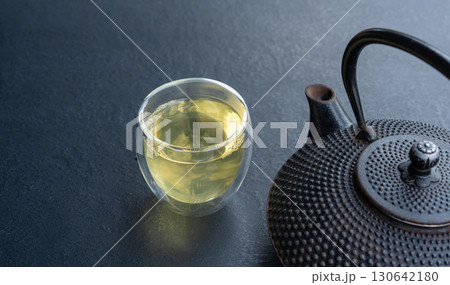 Glass cup with green tea and cast iron teapot on dark table, double wall glass Glass cup with green tea and cast iron teapot on dark table, double wall glass 130642180