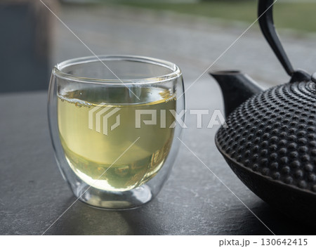 Glass cup with green tea and cast iron teapot on dark table, double wall glass Glass cup with green tea and cast iron teapot on dark table, double wall glass 130642415