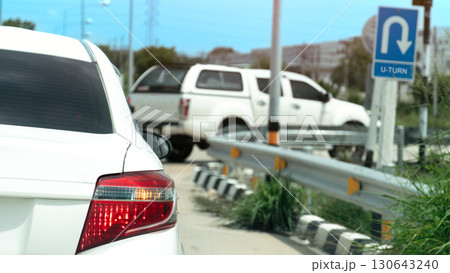 Rear side of white car with right turn signal. Open for u-turn on the road. Front of car the other car waiting for u-turn and sign board for u-turn. Background of city under blue sky. 130643240