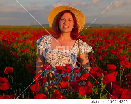 A woman smiles in a straw hat surrounded by red poppies at sunset. Lifestyle, beauty, and natural harmony of rural landscape with human presence and joy. A woman smiles in a straw hat surrounded by red poppies at sunset. Lifestyle, beauty, and natural harmony of rural landscape with human presence and joy. 130644293
